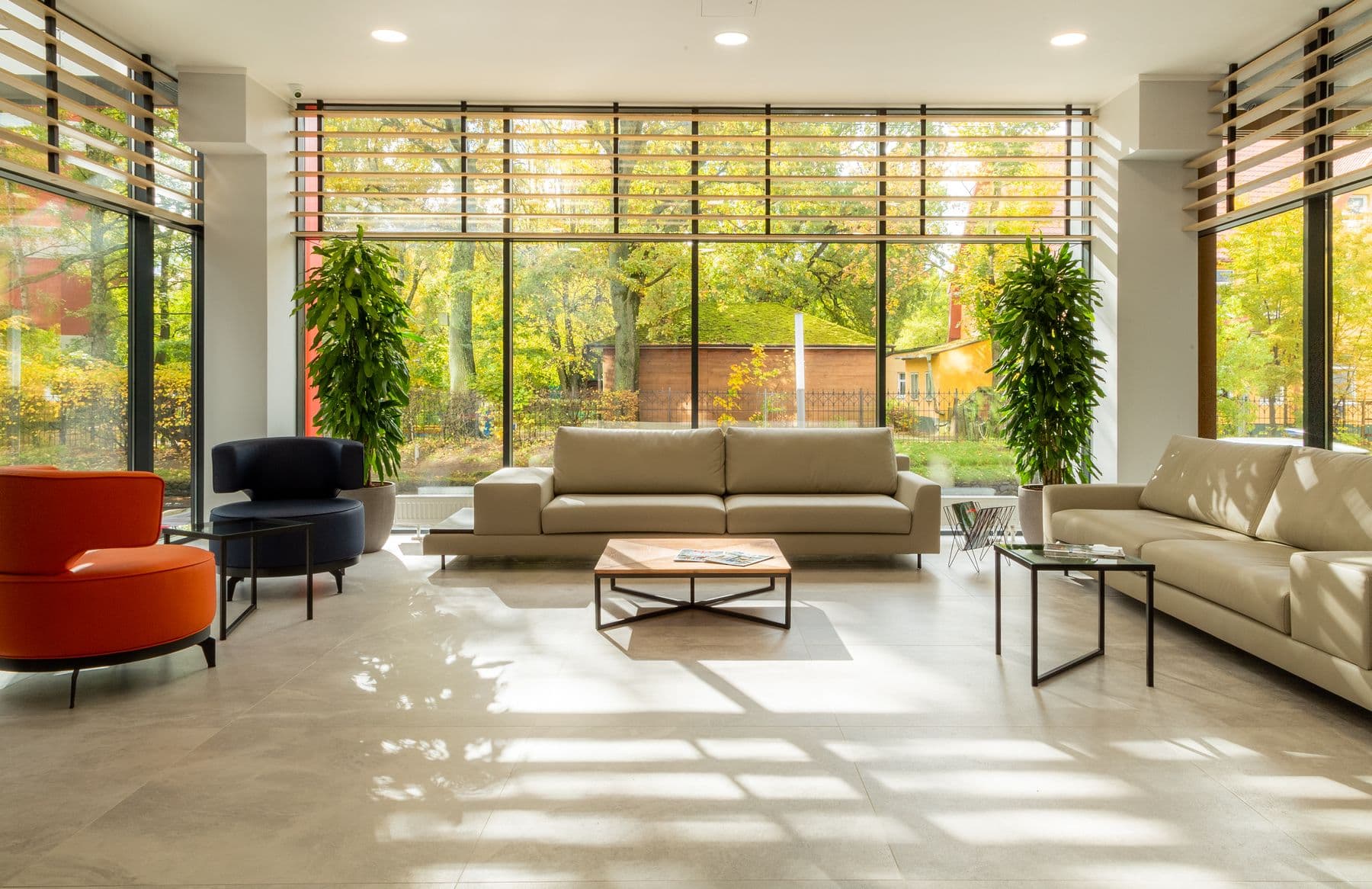 Salón amplio y luminoso con grandes ventanales de suelo a techo, sofás claros y árboles otoñales al otro lado del vidrio